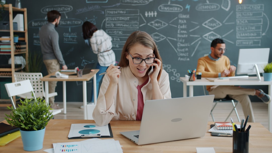 Happy girl blonde is talking on mobile phone showing thumbs-up at desk in coworking workplace while people talking and working with chalkboard in background.